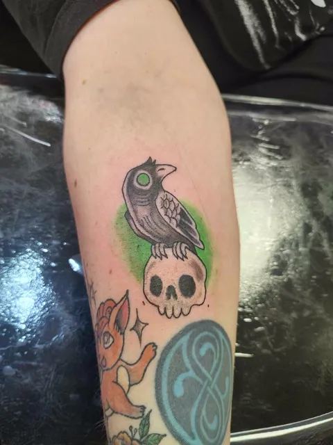 A crow on a skull over a green splash, among other pieces on the forearm, in color