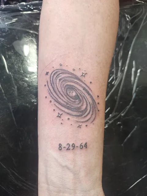 A spiral galaxy above a date on the forearm, black and gray