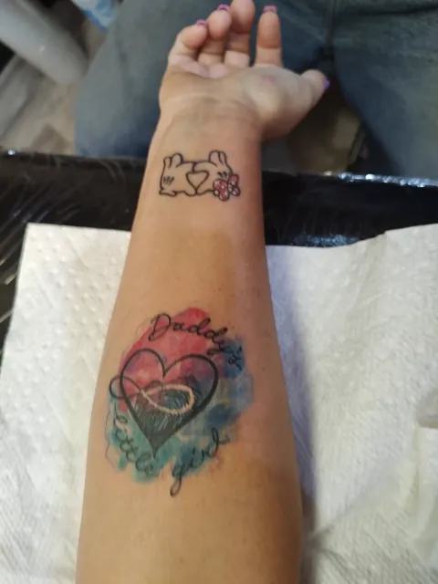 A heart with an infinity sign and Daddy's girl in script over a watercolor splash on the forearm