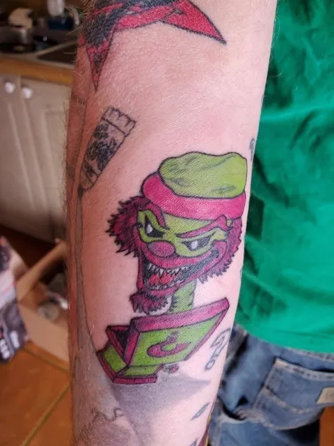 A green-faced character bursting from a box on the forearm, in color