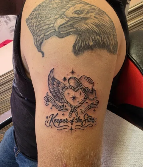 Keeper of the Stars in script under a rope heart with a wing and a cowboy hat, on the upper arm below an eagle, black and gray