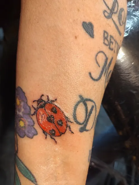 A ladybug beside a purple flower among lettering on the forearm, in color