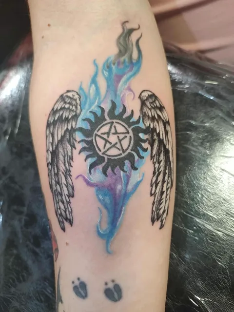 A pentagram sun with angel wings and blue flames on the forearm, in color