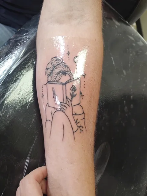 A woman reading a book with a dagger and a rose, under a crescent moon, on the forearm, fine line
