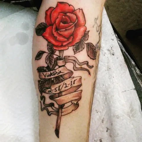 A red rose over a ribbon banner on the forearm, in color