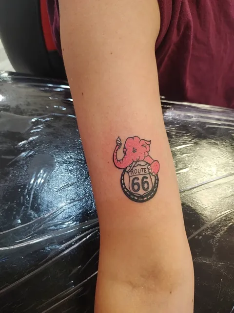 A small pink elephant on a Route 66 badge on the forearm, in color