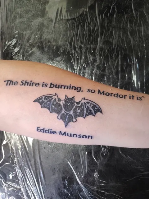 A bat between the words The Shire is burning, so Mordor it is and the name Eddie Munson, on the forearm