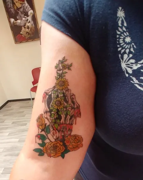 A skeleton hand holding sunflowers and roses on the upper arm, in color