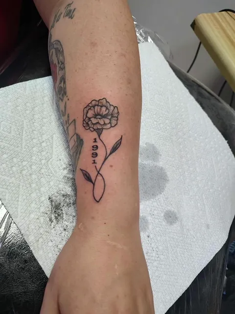 One small rose on a stem with a year, on the forearm, fine line